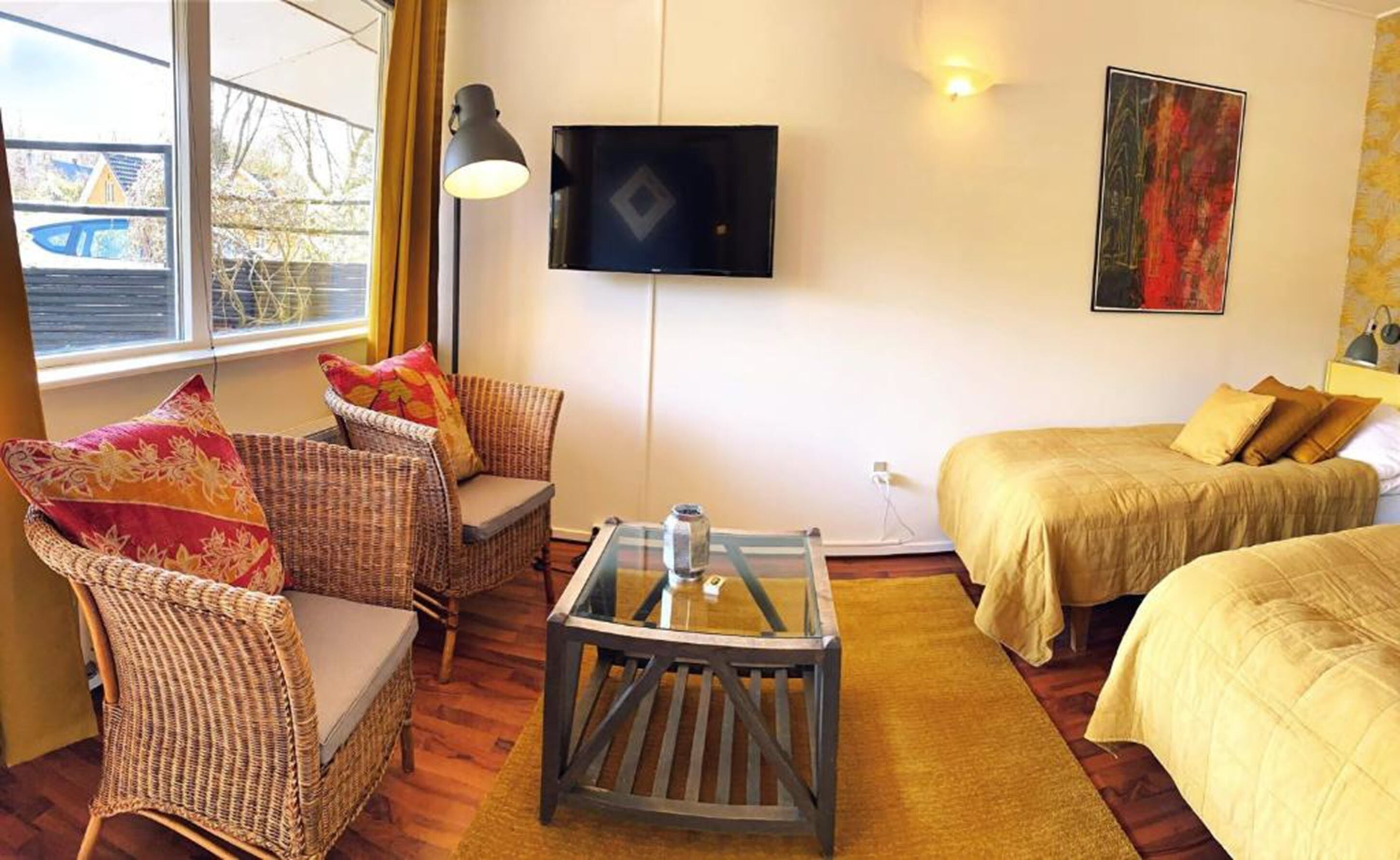 Une chambre meublée avec des fauteuils en osier, une table basse avec un plateau en verre, un lit avec une literie jaune, un téléviseur fixé au mur et un tableau accroché au mur.