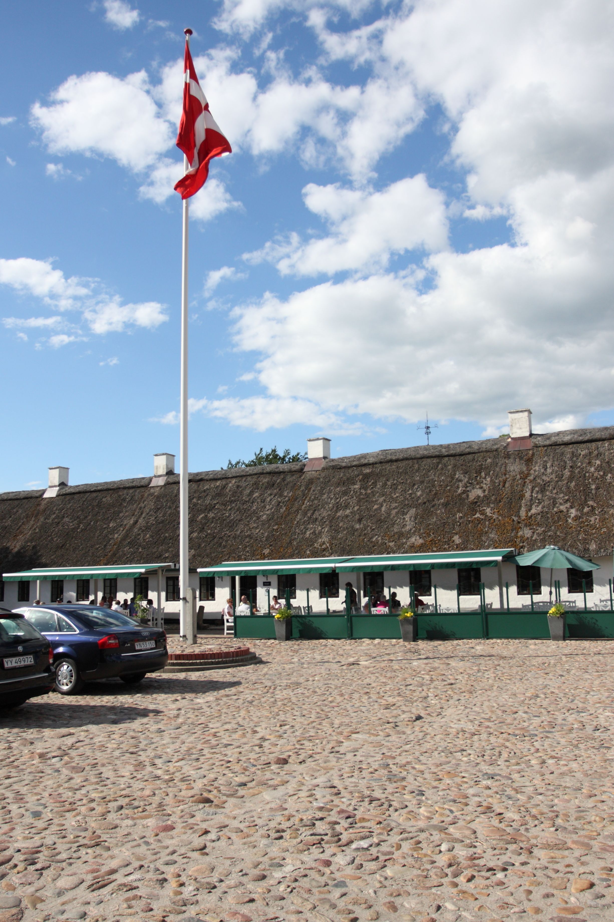 A building with a thatched roof, a flagpole with a flag, and a cobblestone ground.