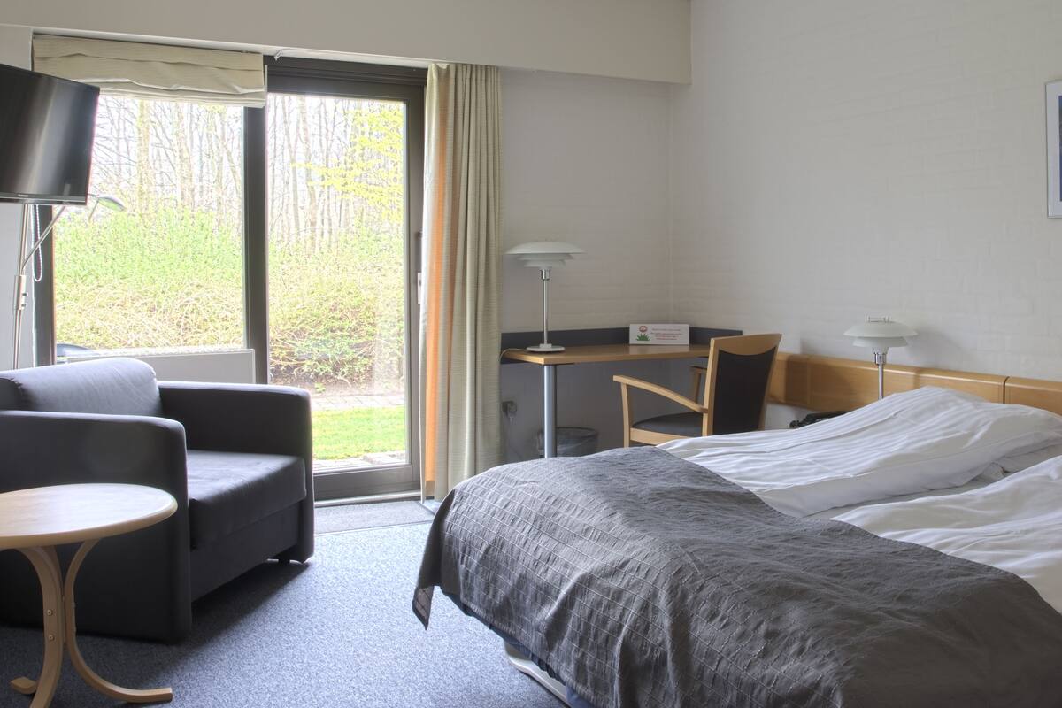 Une chambre d’hôtel équipée d’un lit, d’un bureau, d’une chaise, d’une télévision et d’une fenêtre donnant sur la verdure.