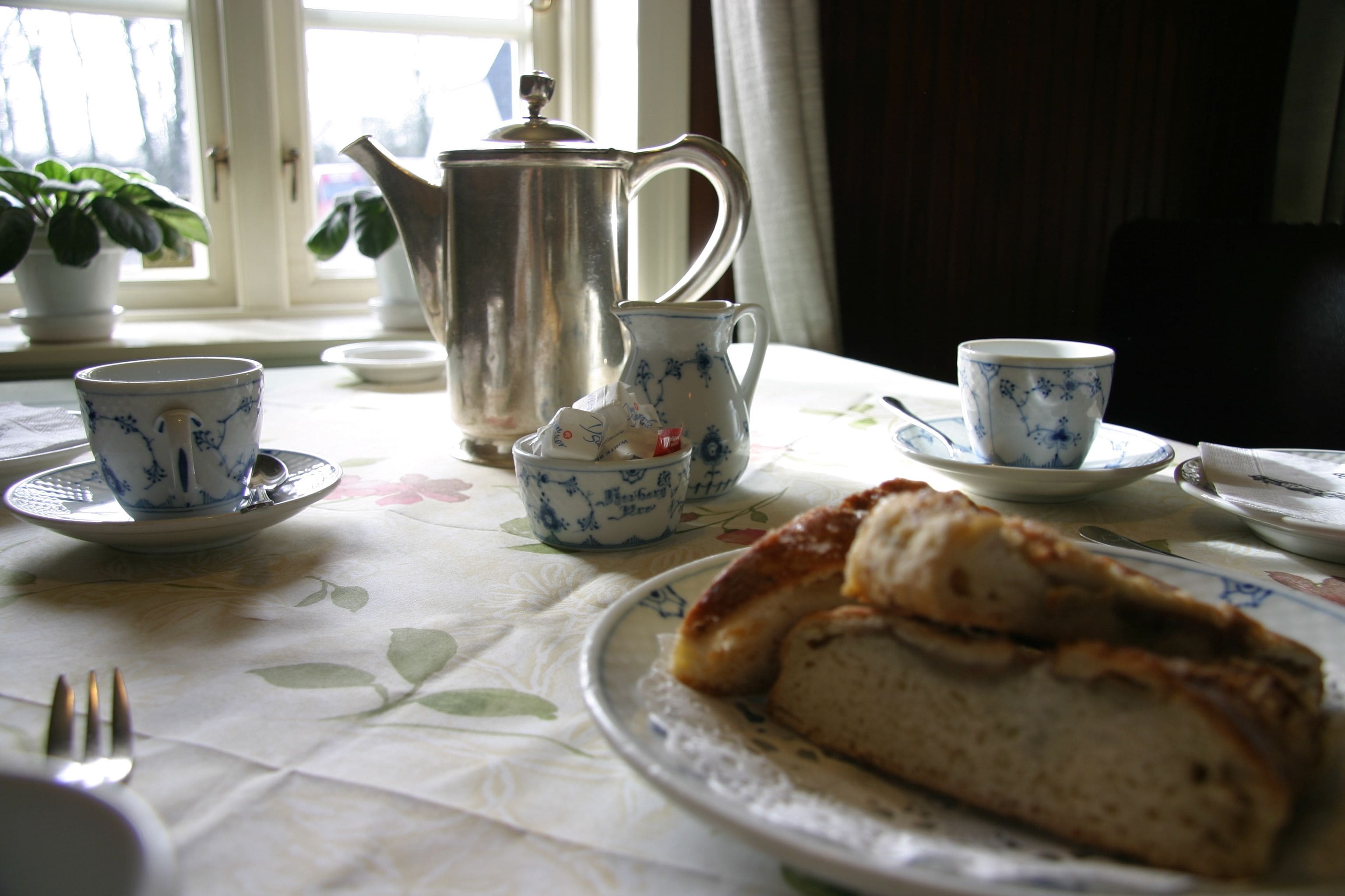 Una mesa con mantel floral, una tetera, tazas, platillos y un plato con un pastel.