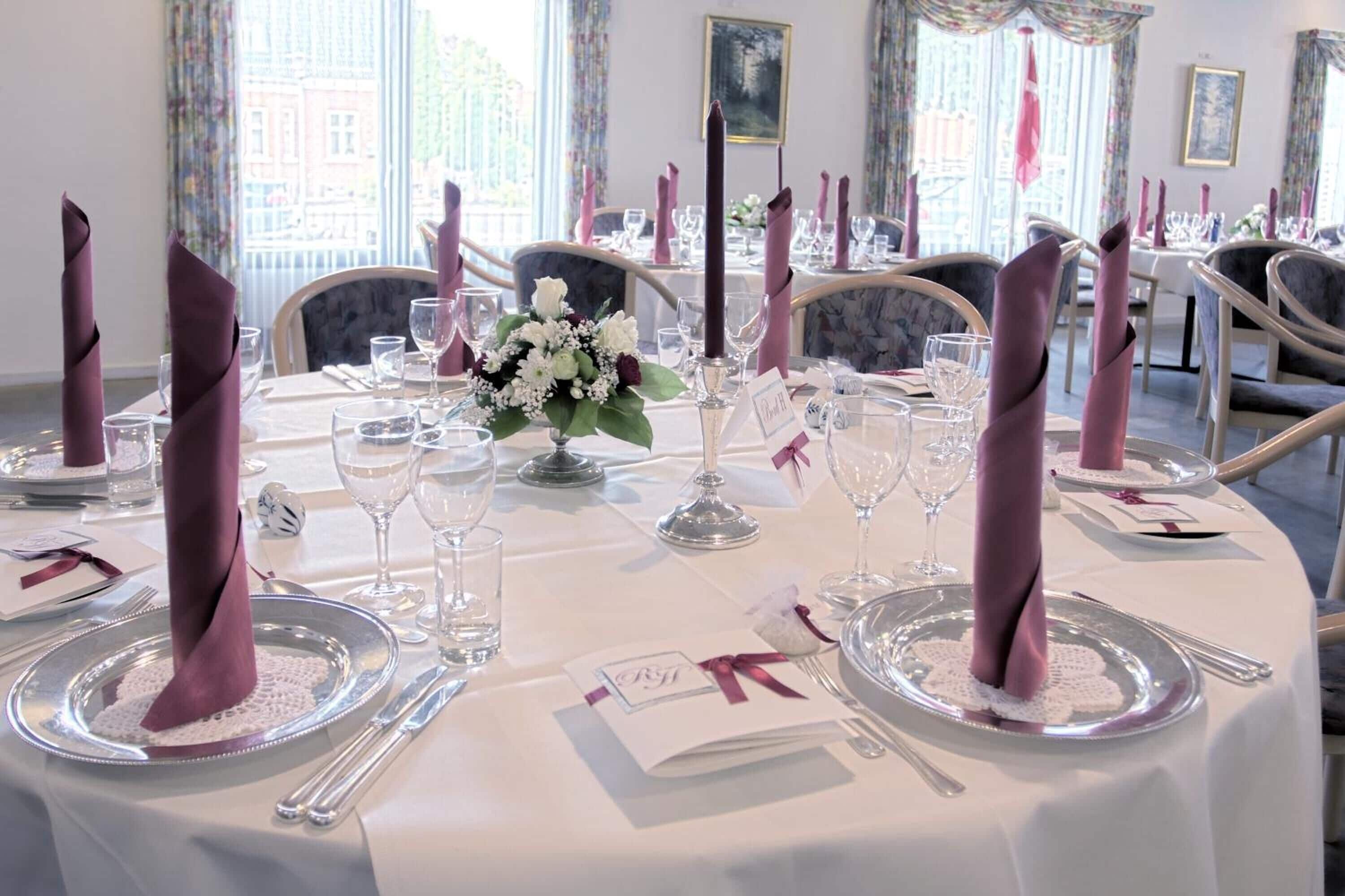 A formal dining room with round tables set for a meal, each with a white tablecloth, silverware, and pink napkins.