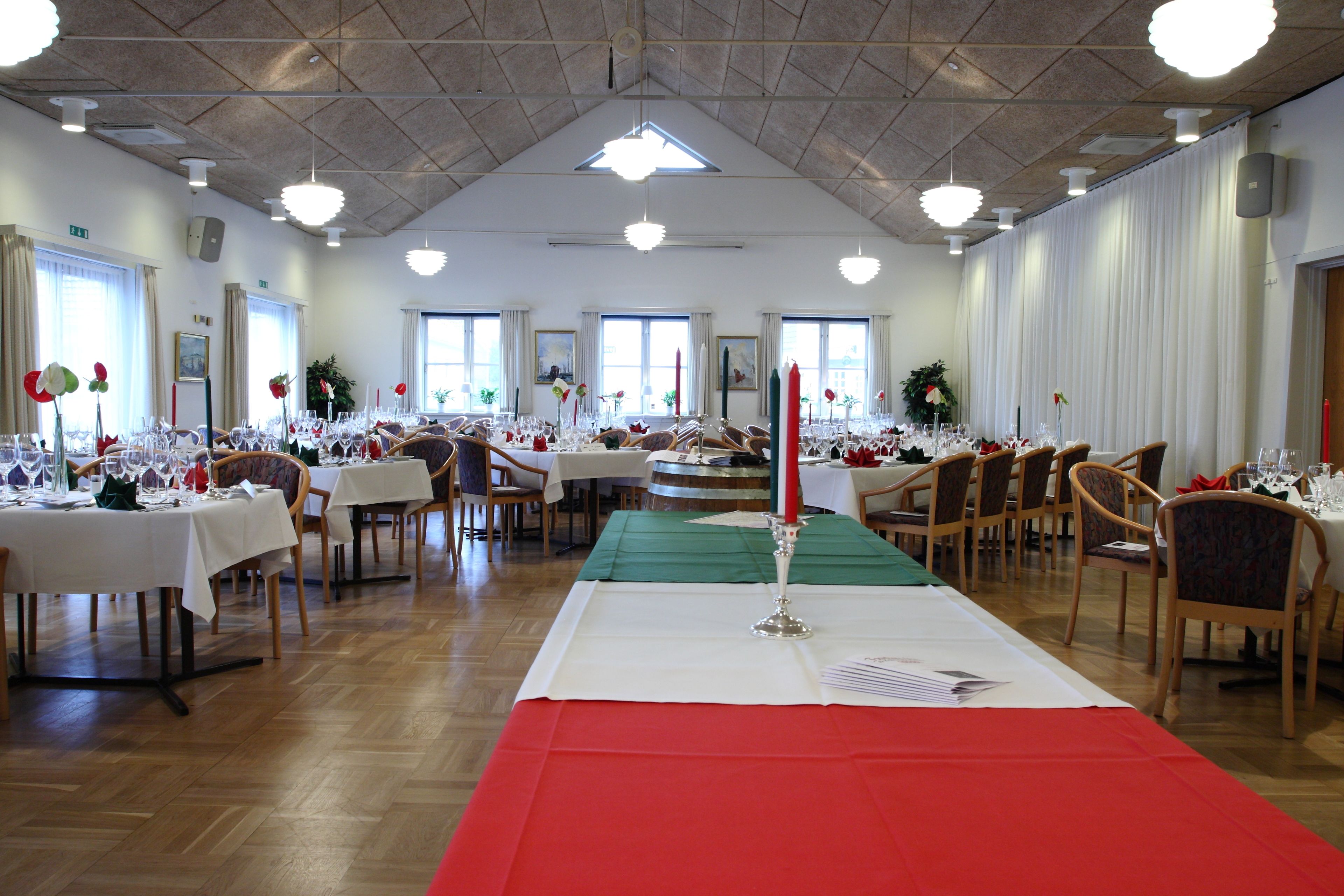 A spacious banquet hall with multiple tables set for dining, large windows, and a high ceiling with hanging lights.