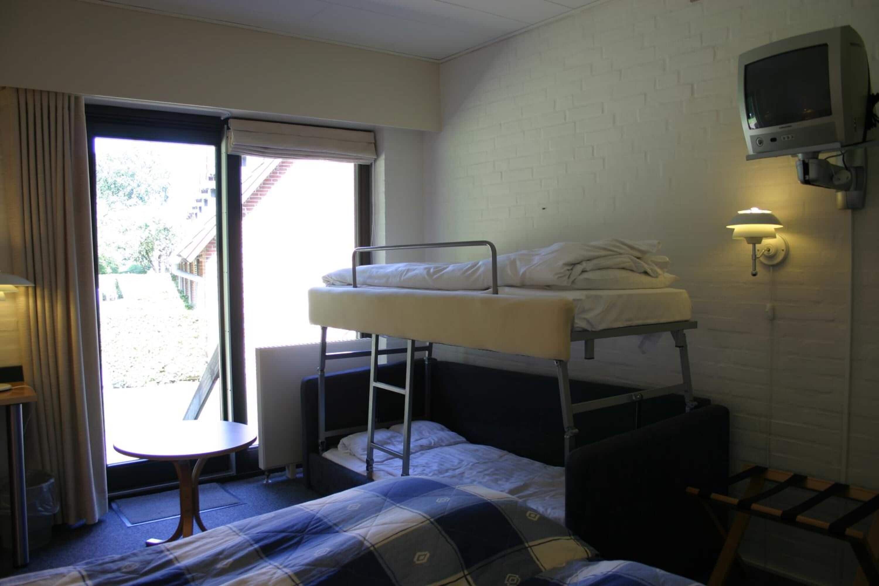 Een slaapzaal met stapelbedden, een kleine tafel, een stoel, een aan de muur bevestigde televisie en een raam met gordijnen.