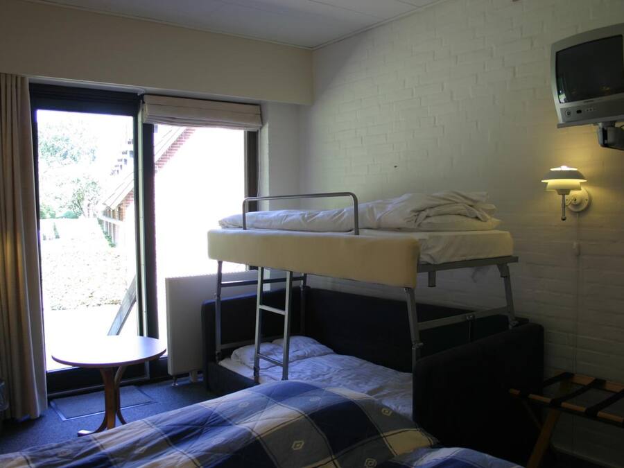 Quarto em dormitório com beliches, uma mesa pequena, uma cadeira, televisão fixada na parede e uma janela com cortinas.