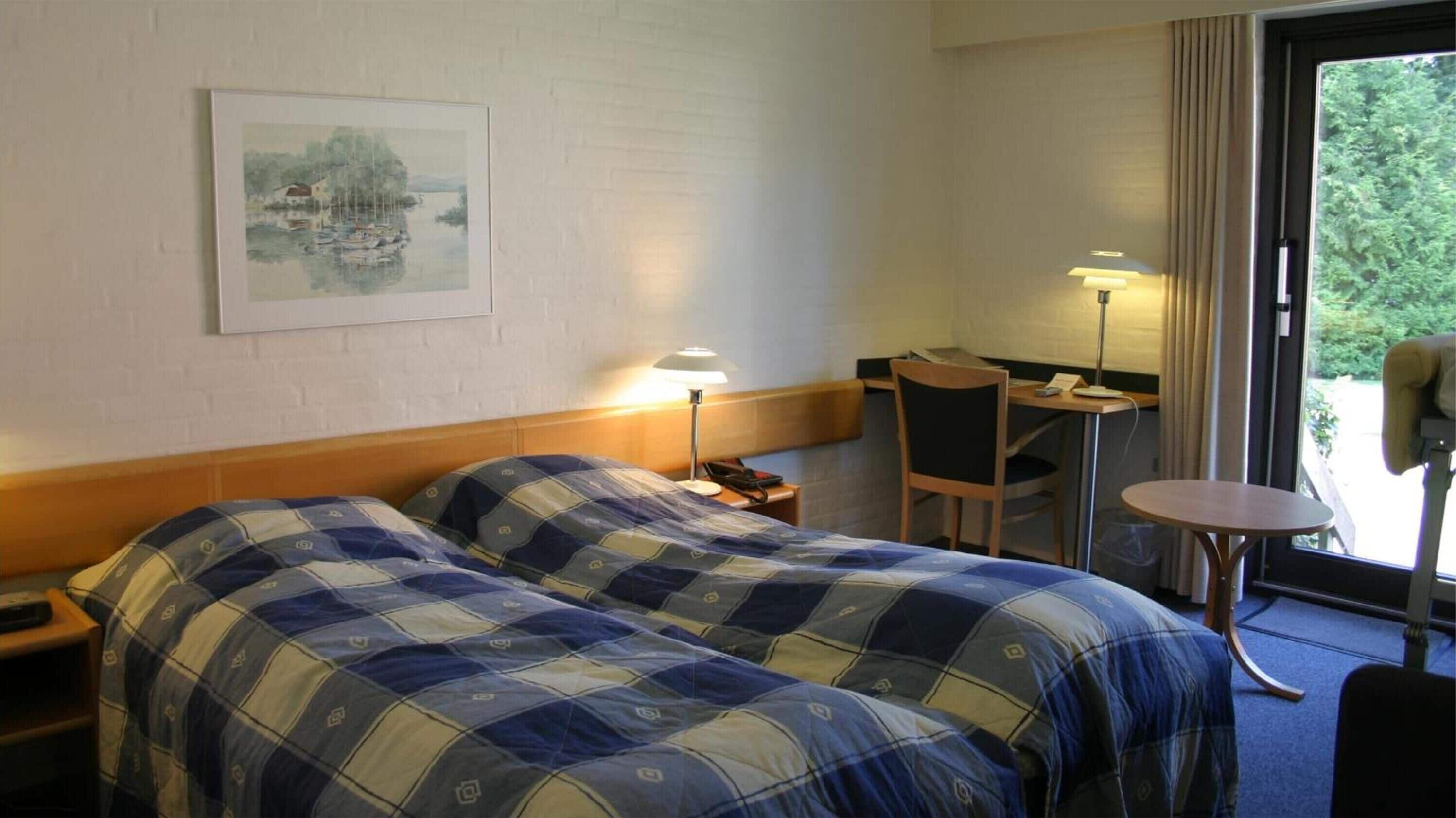A hotel room with two beds, a desk, a chair, a lamp, and a painting on the wall.