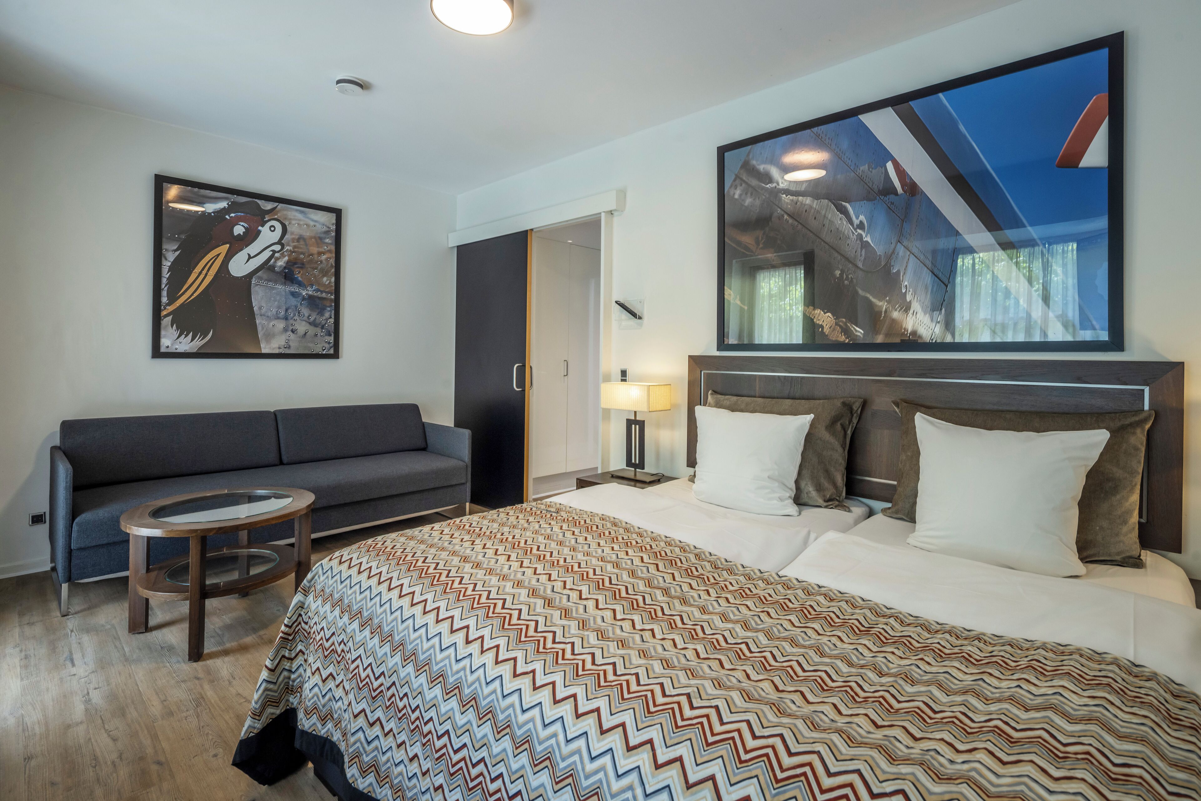 Ein modernes Hotelzimmer mit einem großen Bett, einer Couch, einem kleinen runden Tisch und Kunstwerken an den Wänden.