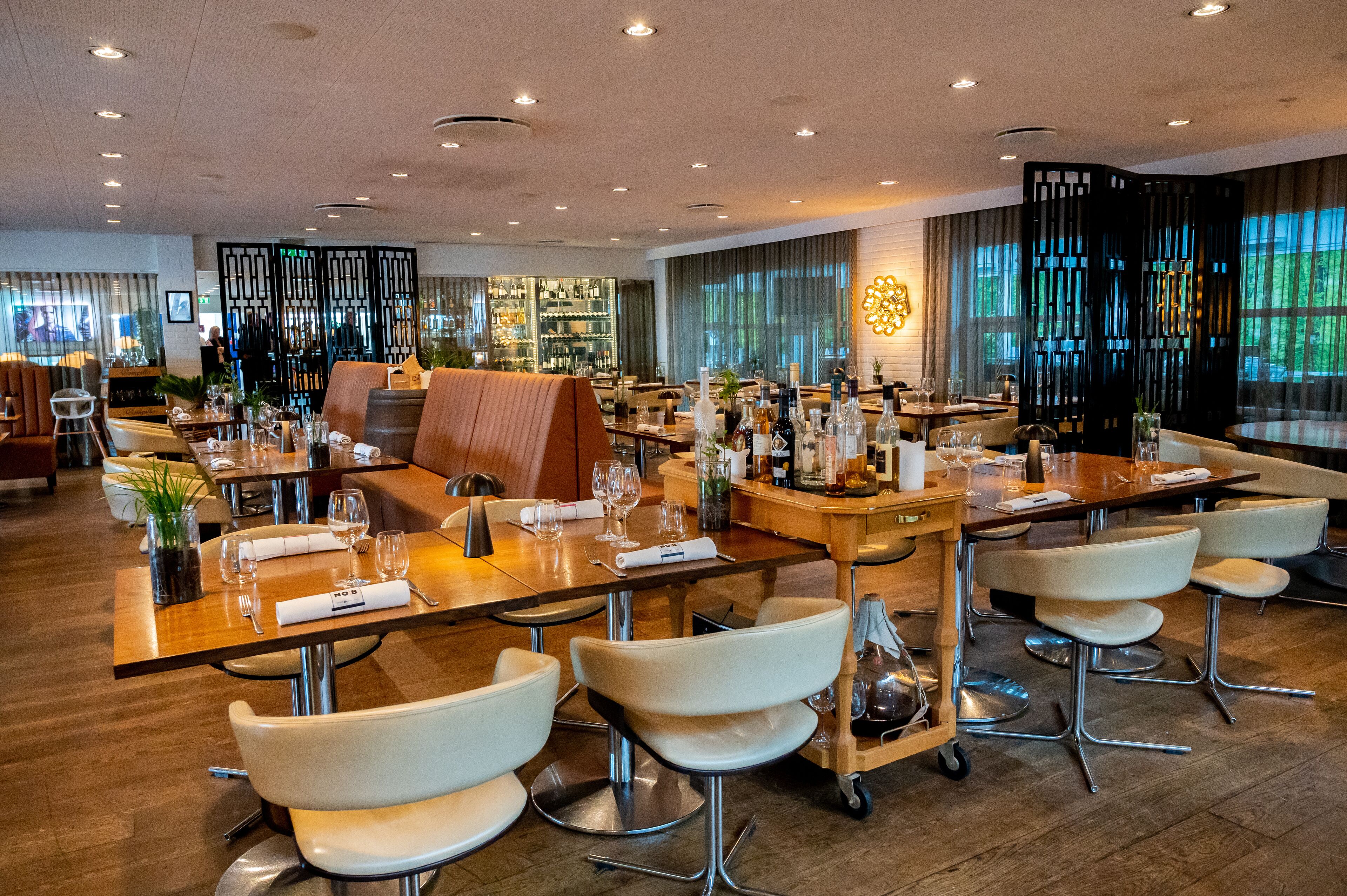 A modern restaurant with wooden tables, white chairs, and a bar area.