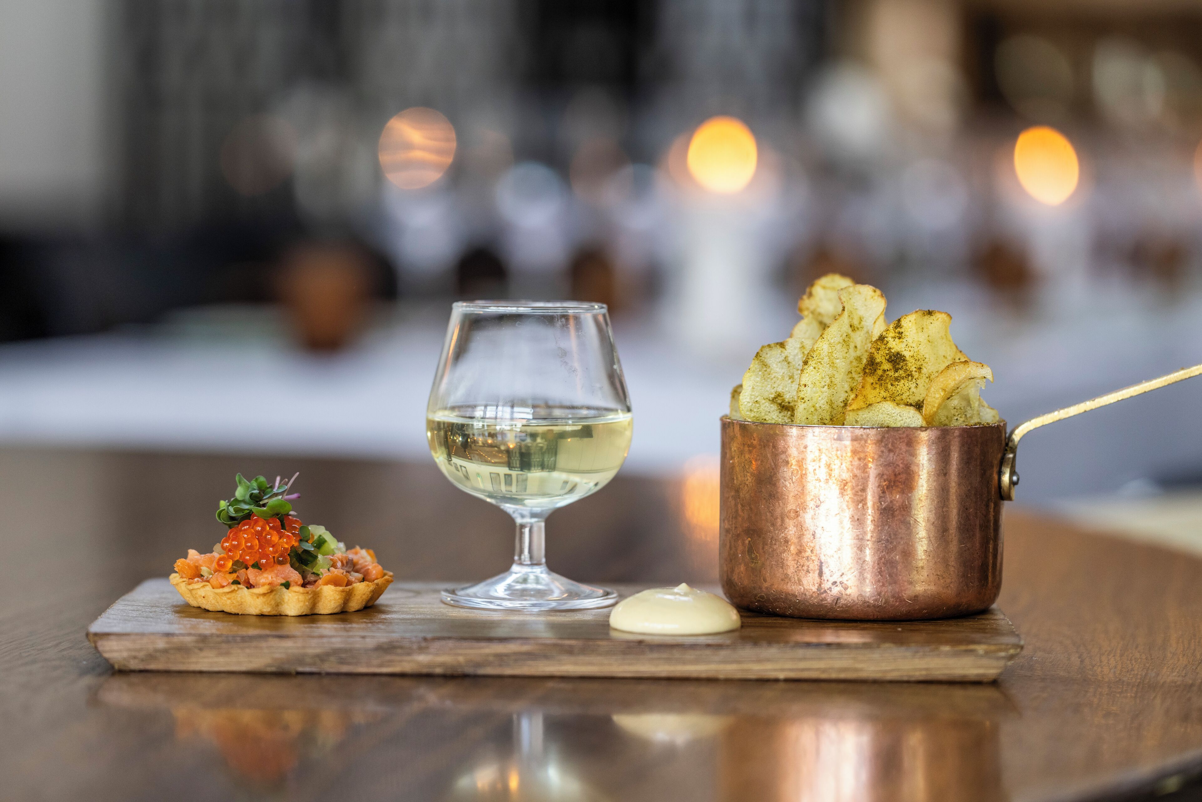 A dessert plate with a tart, a glass of white wine, and a copper pot with fried items on a wooden table.