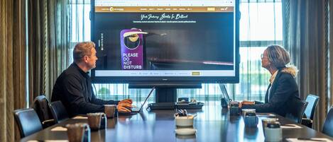 Una sala de conferencias con una pantalla grande mostrando la página web de un hotel. Dos personas están sentadas en una mesa, con tazas de café y portátiles.