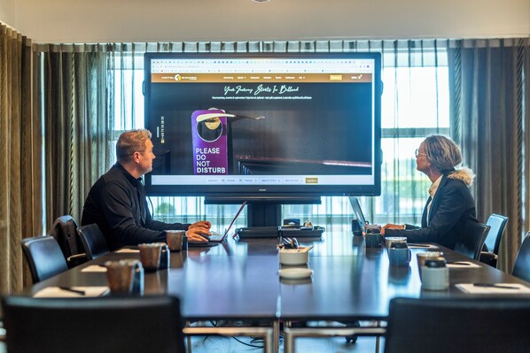 A conference room with a large screen displaying a hotel website. There are two people seated at a table with coffee cups and laptops.