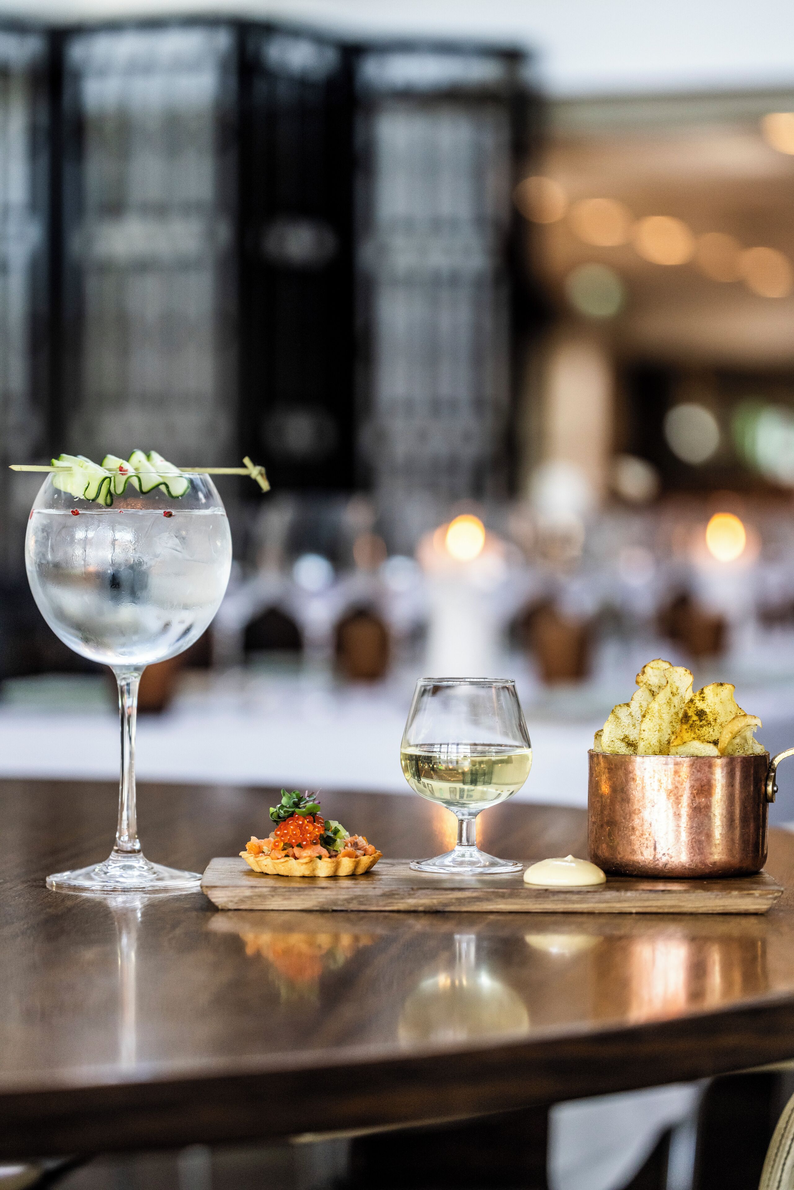 A table with a glass of water, a small dish of food, a glass of white wine, and a copper pot of chips.