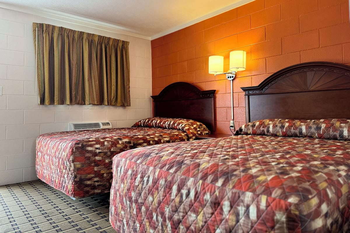 A hotel room with two beds, a brick wall, a window with curtains, and a wall-mounted lamp.