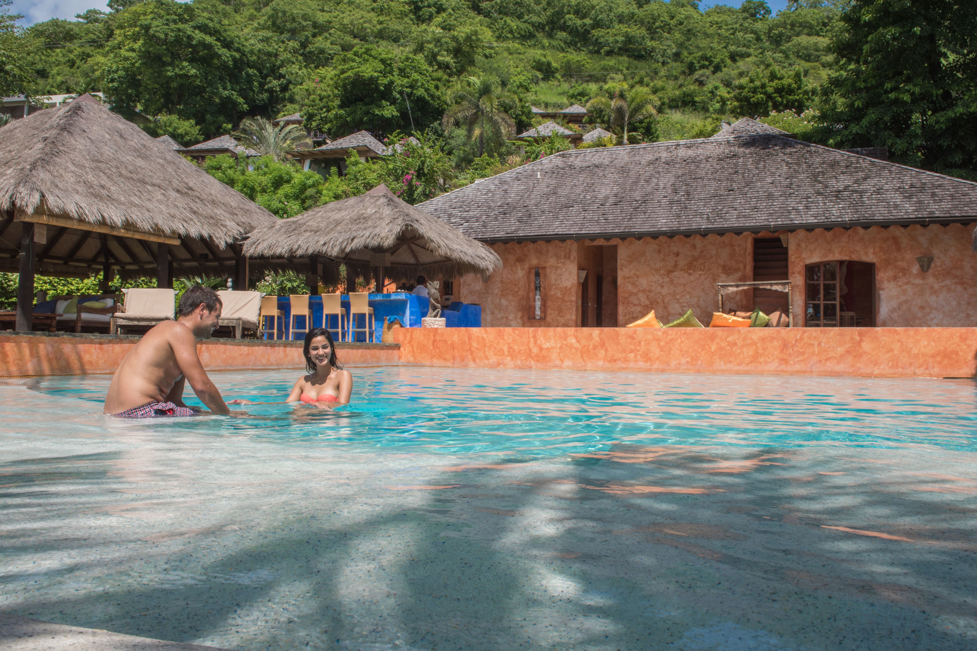 Una piscina con una pareja, un edificio con techo de paja y exuberante vegetación al fondo.