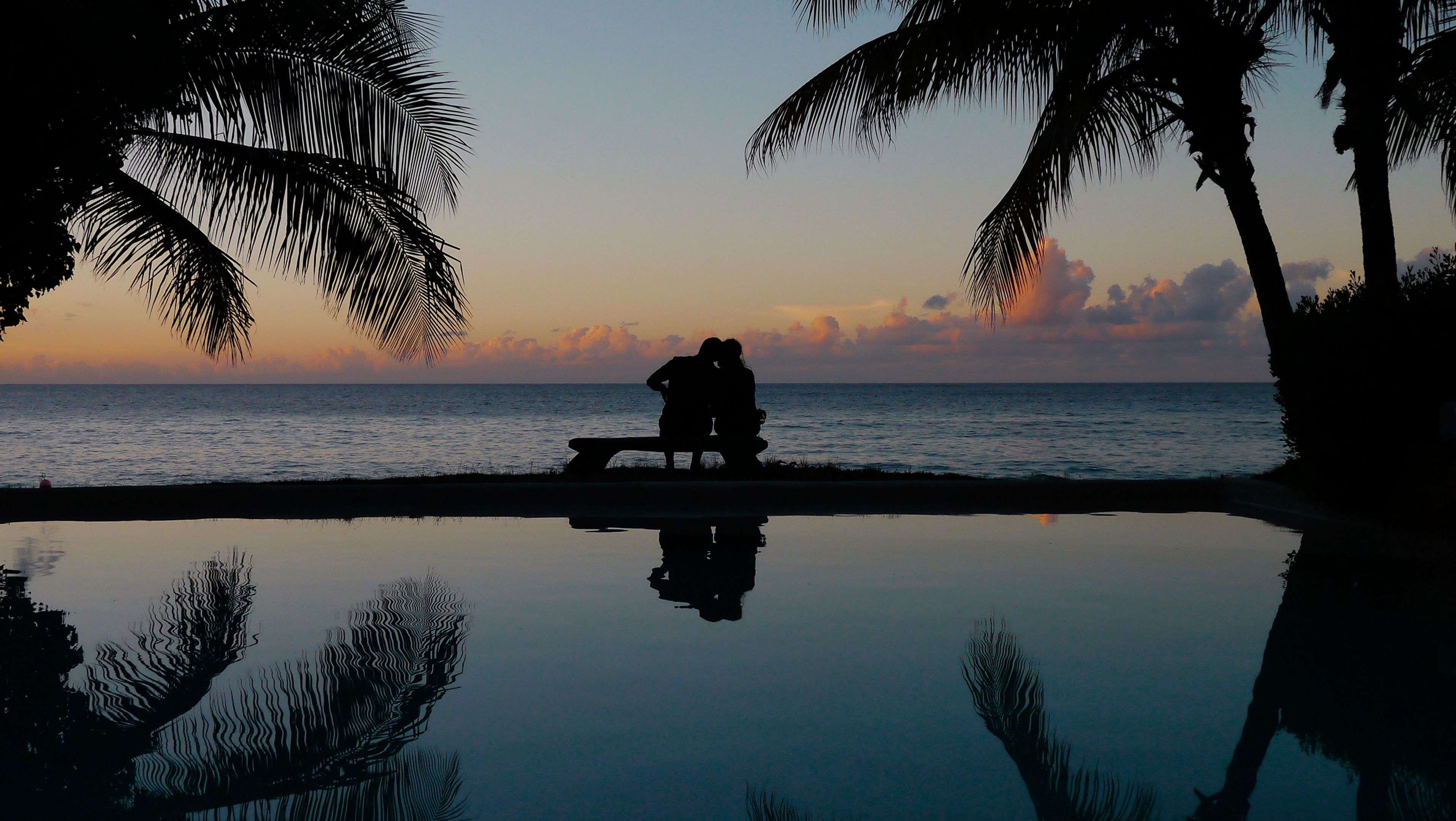 A couple sitting on a bench by a reflective pool with palm trees and a sunset view.