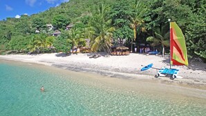 Une plage avec une eau turquoise et limpide, un rivage sablonneux et quelques personnes qui se baignent. On y trouve des palmiers et quelques parasols de plage.