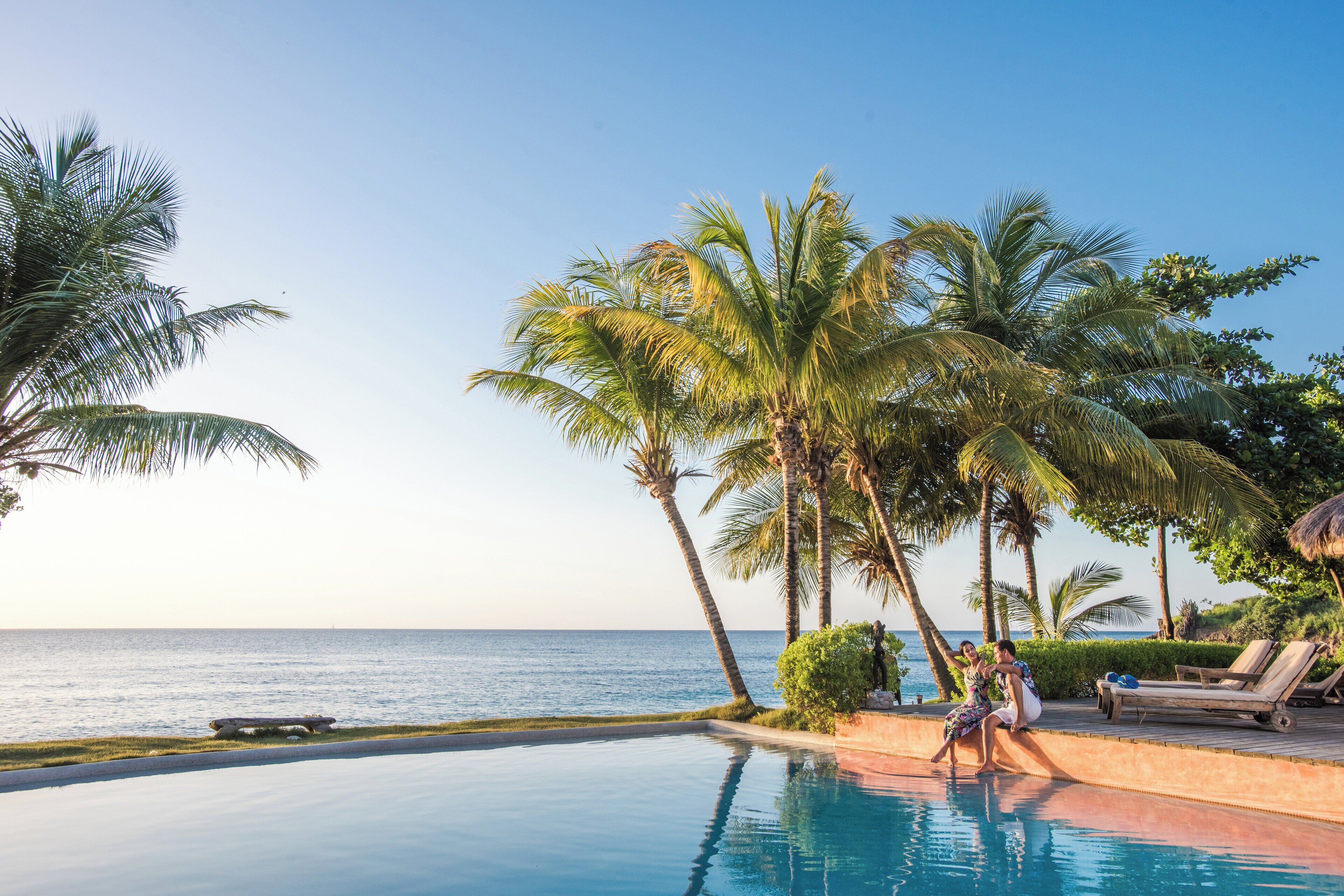 A poolside area with a view of the ocean, palm trees, and lounge chairs.