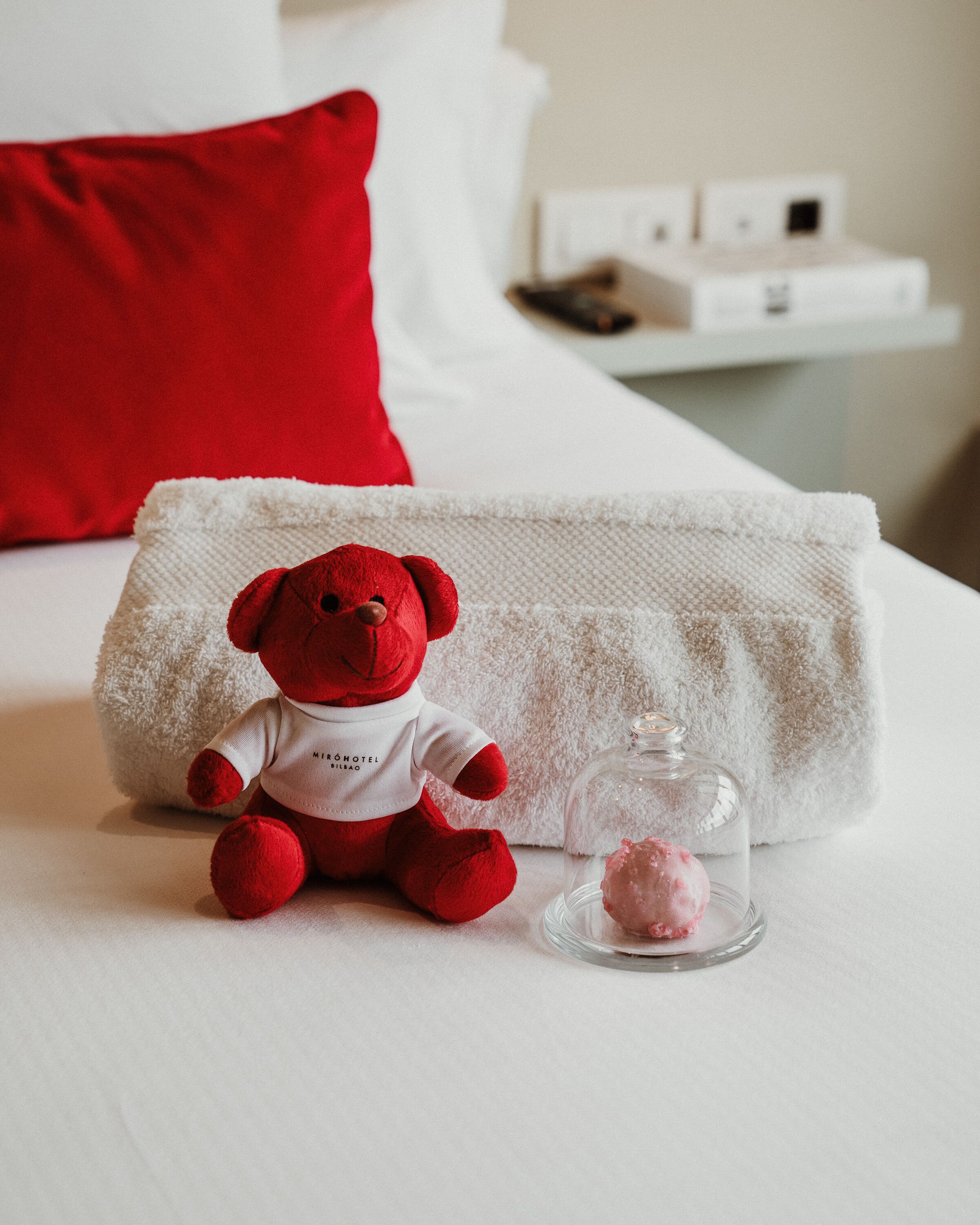 Un osito rojo sobre una cama blanca con un cojín rojo y un frasco con un líquido rosado.