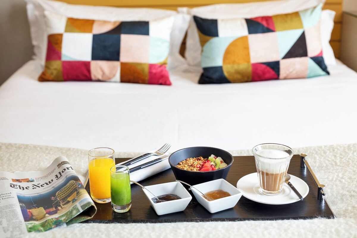A neatly made breakfast tray with a newspaper, a glass of orange juice, a bowl of cereal with fruits, a glass of milk, and a cup of coffee on a bed with colorful pillows.