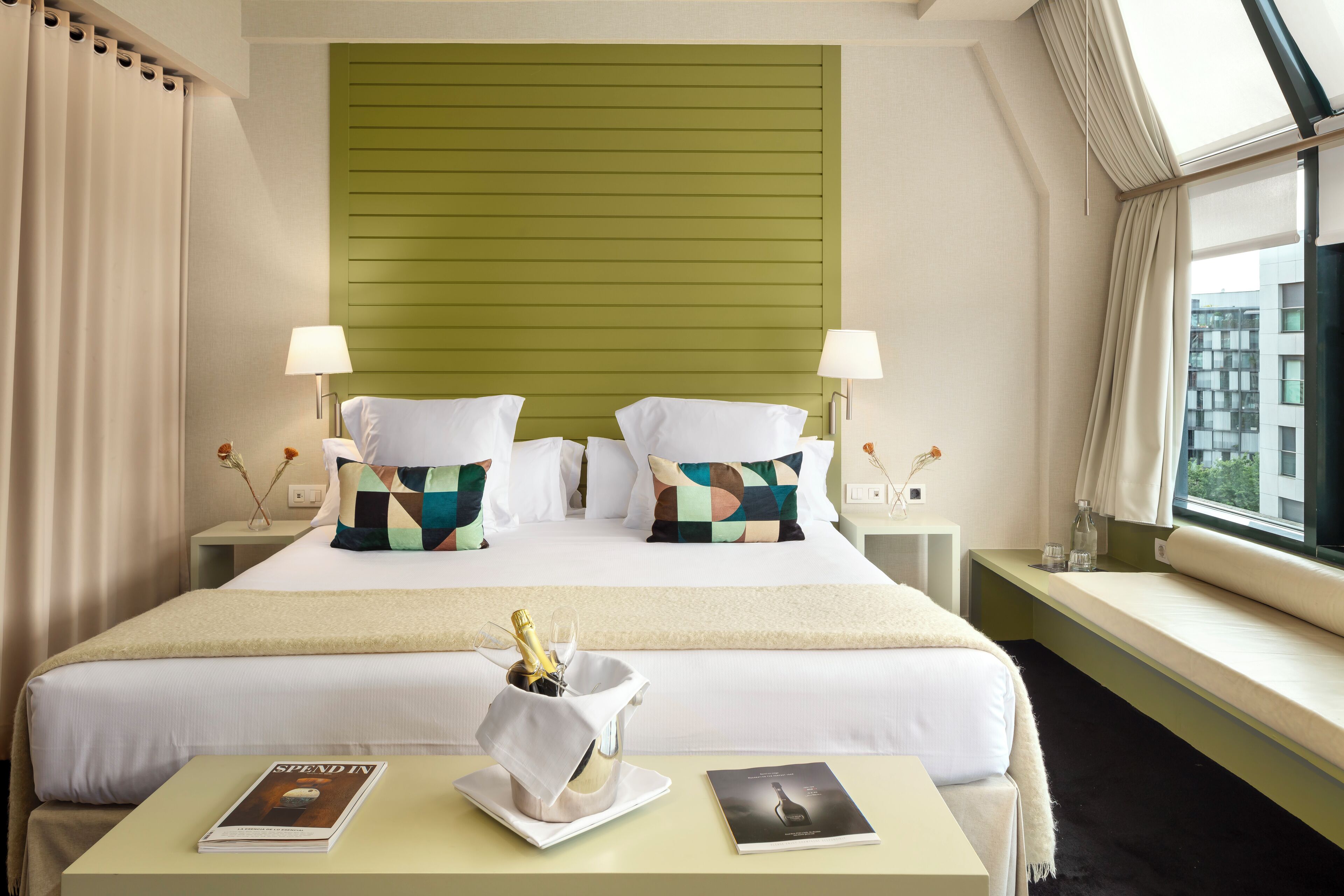 A hotel room with a large bed, two bedside tables, a window with a view, and a small table with a magazine and a bottle of champagne.