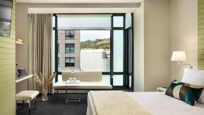 A hotel room with a bed, a desk, a chair, a window with a view of buildings, and a lamp. - Hotel Miro (Bilbao)