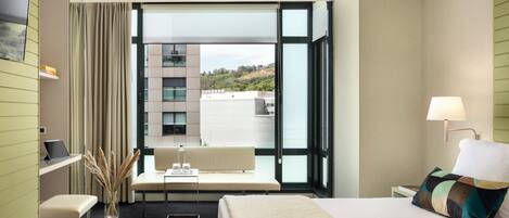 A hotel room with a large window, a bed, a desk, and a chair.