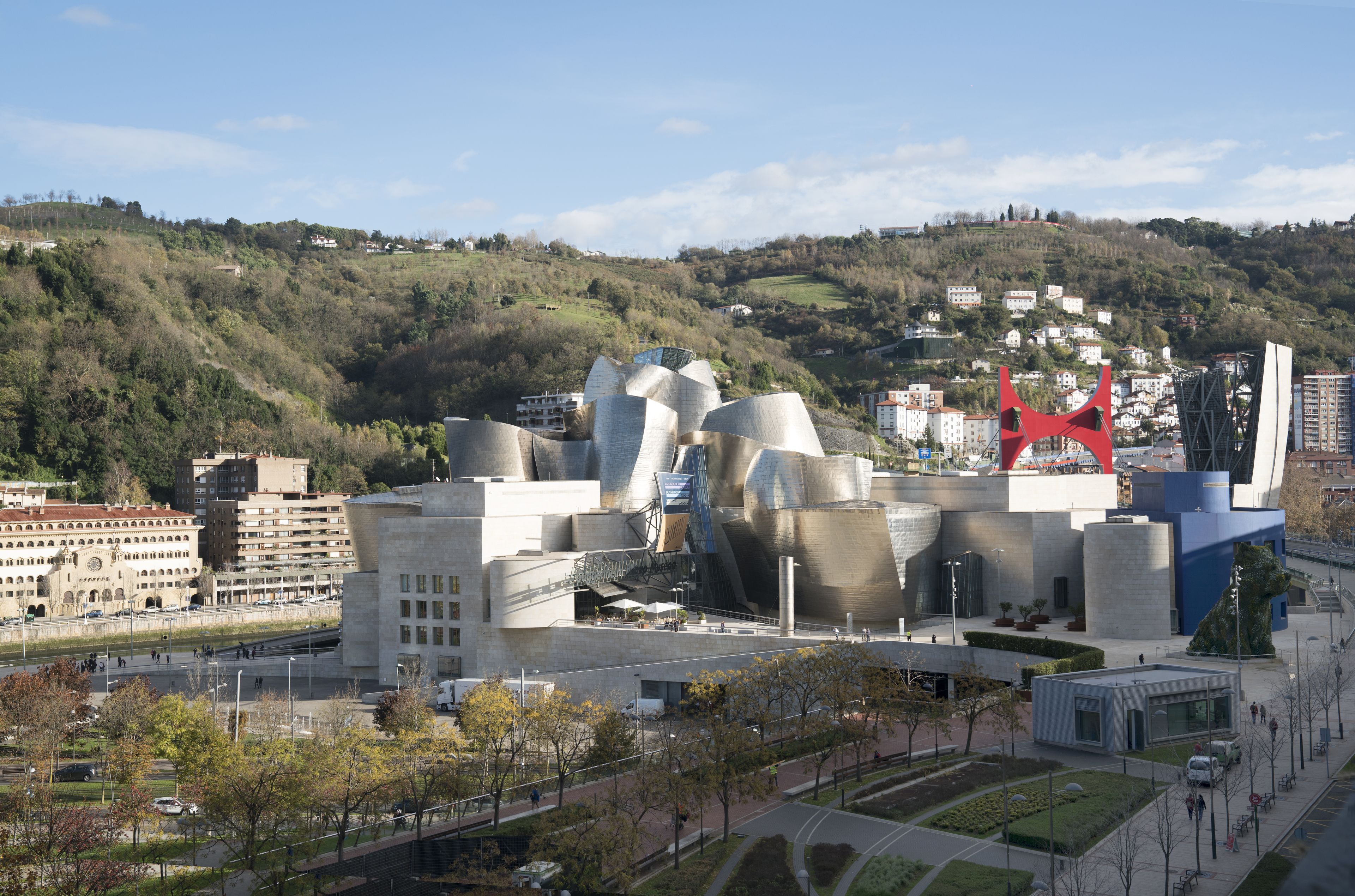 Un edificio moderno con un diseño arquitectónico singular, rodeado de un área ajardinada con árboles y vistas a un paisaje montañoso en el fondo.