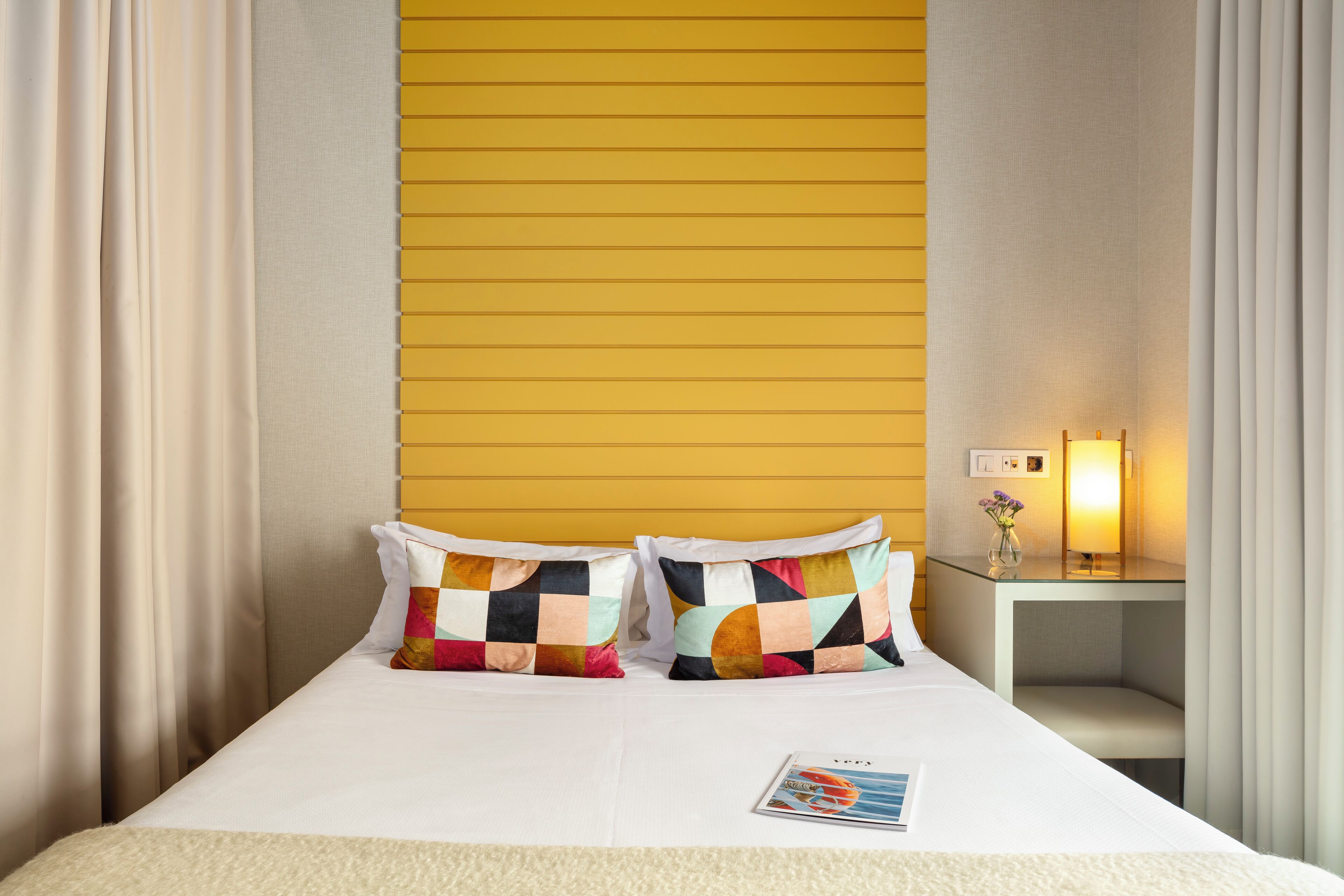 Habitación de hotel con una cama, dos almohadas coloridas, una mesita de noche con una lámpara y una revista sobre la cama.