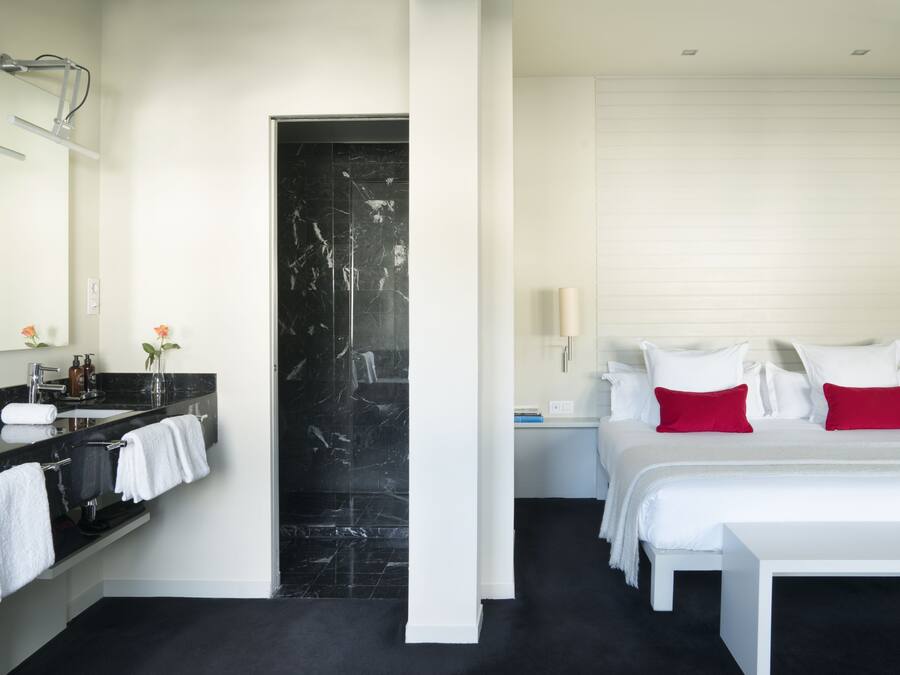 A modern hotel room with a black marble bathroom, white bed with red pillows, and a large mirror above the sink.