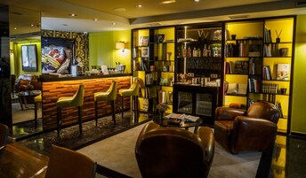 Zona de bar con mostrador de madera y sillas amarillas, estantería con libros y objetos decorativos, y asientos de cuero al frente.