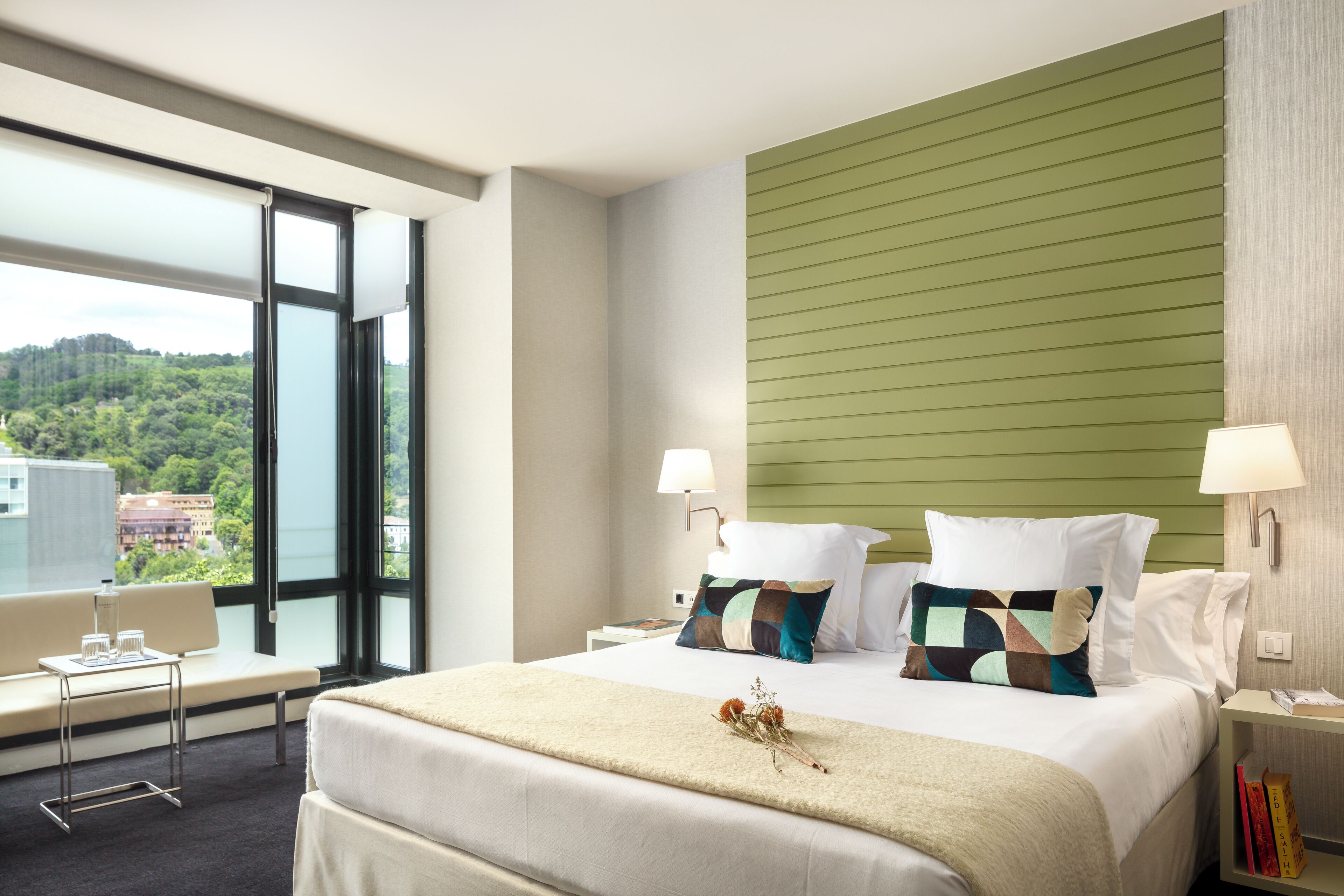 Une chambre d’hôtel moderne avec un grand lit, deux lampes de chevet, une vue sur le paysage urbain et un mur d’accent vert.
