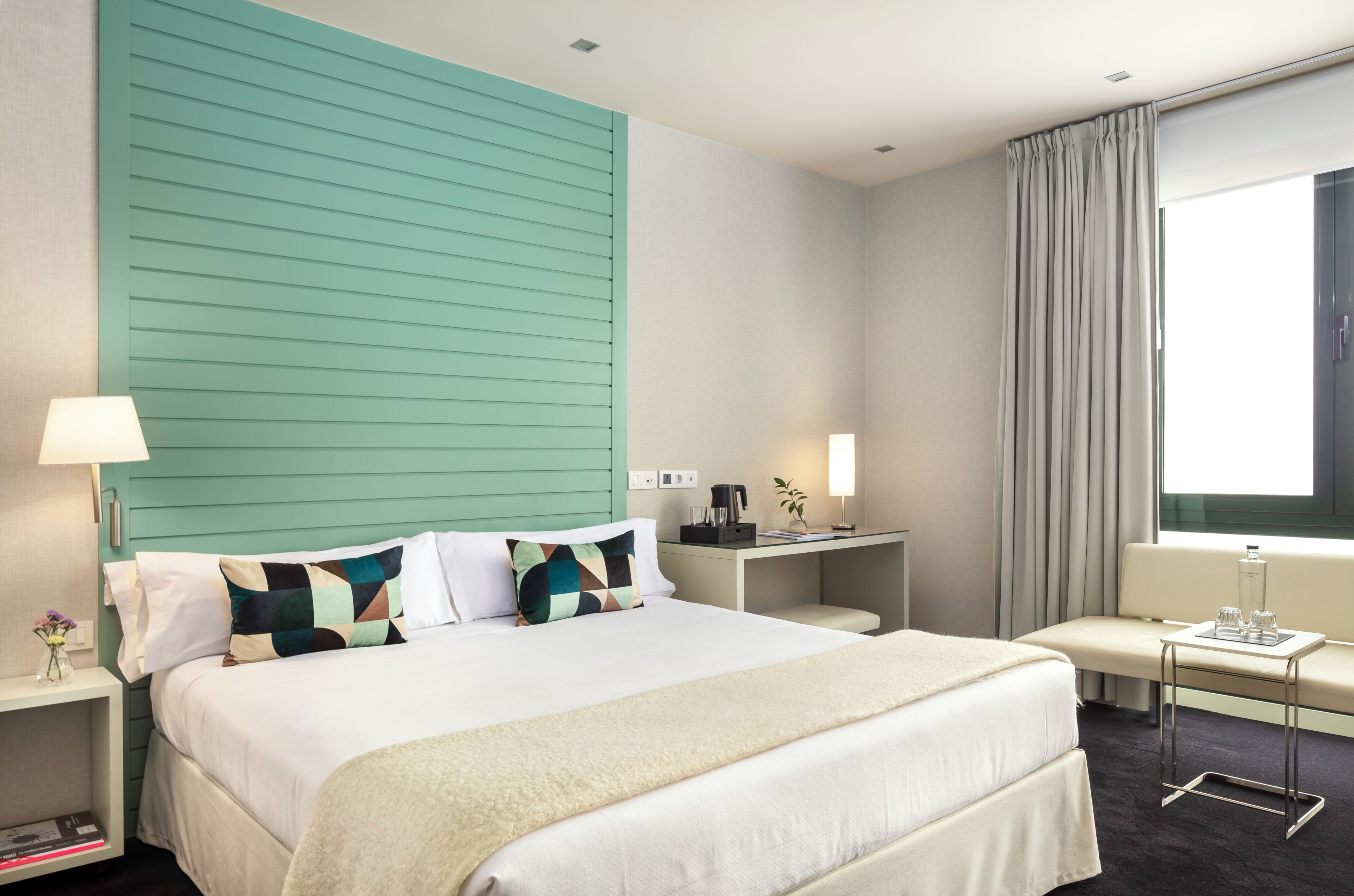 Habitación de hotel con cama, mesita de noche, lámpara y ventana con cortinas.