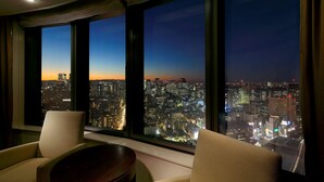 Una habitación con un amplio ventanal que ofrece vistas panorámicas de la ciudad al anochecer. Se ven dos sillas y una mesita en primer plano.