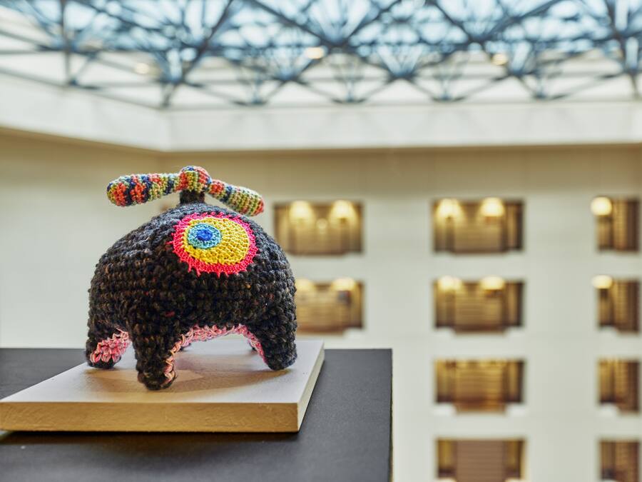 A crocheted elephant sculpture on a wooden platform in a building with a glass ceiling and multiple windows.