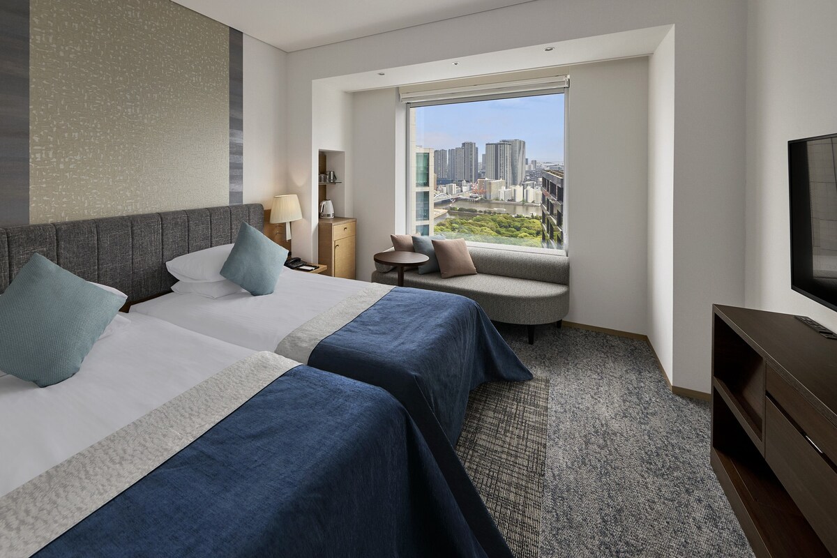 A hotel room with two beds, a sofa, a TV, and a window with a city view.