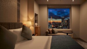 Habitación de hotel con cama, una mesita, una lámpara y vista a un paisaje urbano de noche.