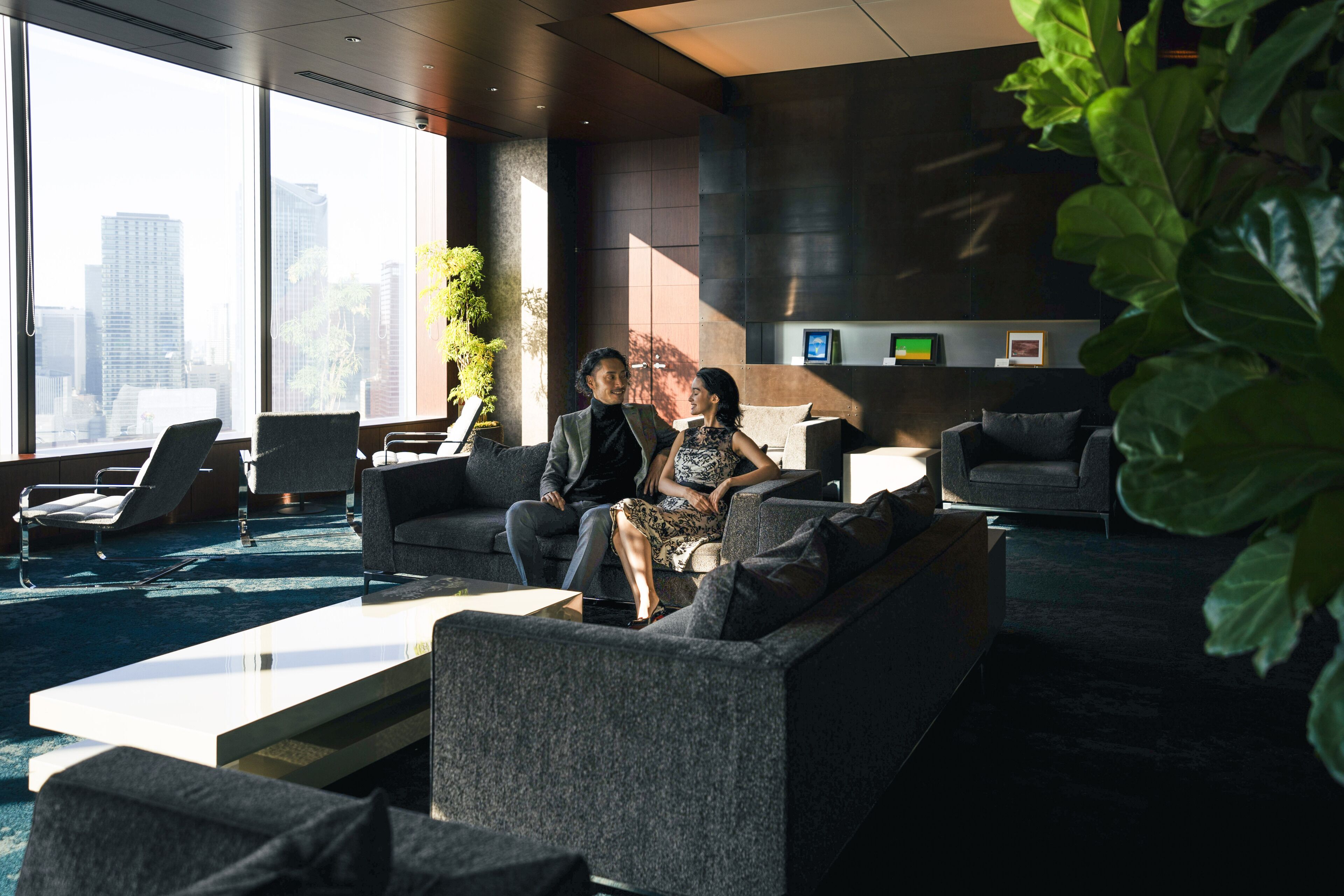 A modern lounge area with a city view, comfortable seating, and a large plant.