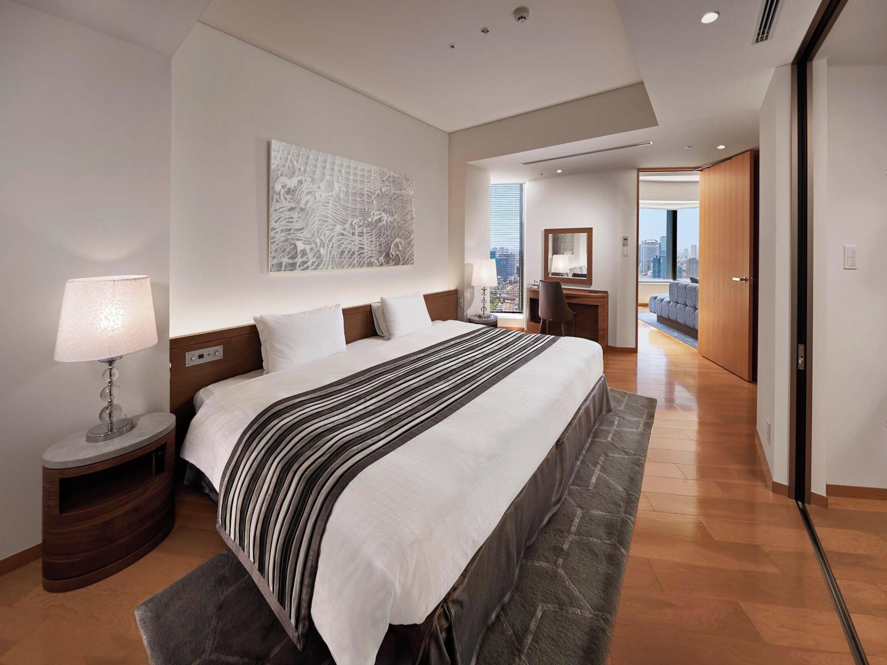 Ein modernes Hotelzimmer mit einem großen Bett, einem Schreibtisch, einem Stuhl, einer Lampe, Blick auf die Stadt und einer Tür zu einem weiteren Raum.