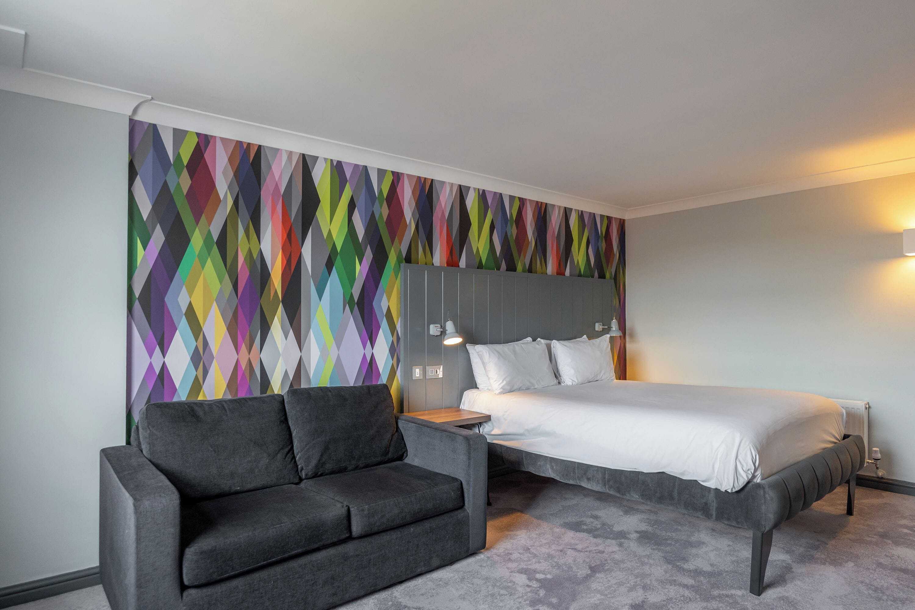 Une chambre d’hôtel moderne avec un lit, un canapé gris et un motif géométrique coloré sur le mur.