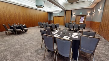 Un salón de conferencias con mesas y sillas redondas, suelo alfombrado, paneles de madera y luces de techo.