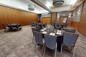 Un salón de conferencias con mesas y sillas redondas, suelo alfombrado, paneles de madera y luces de techo.