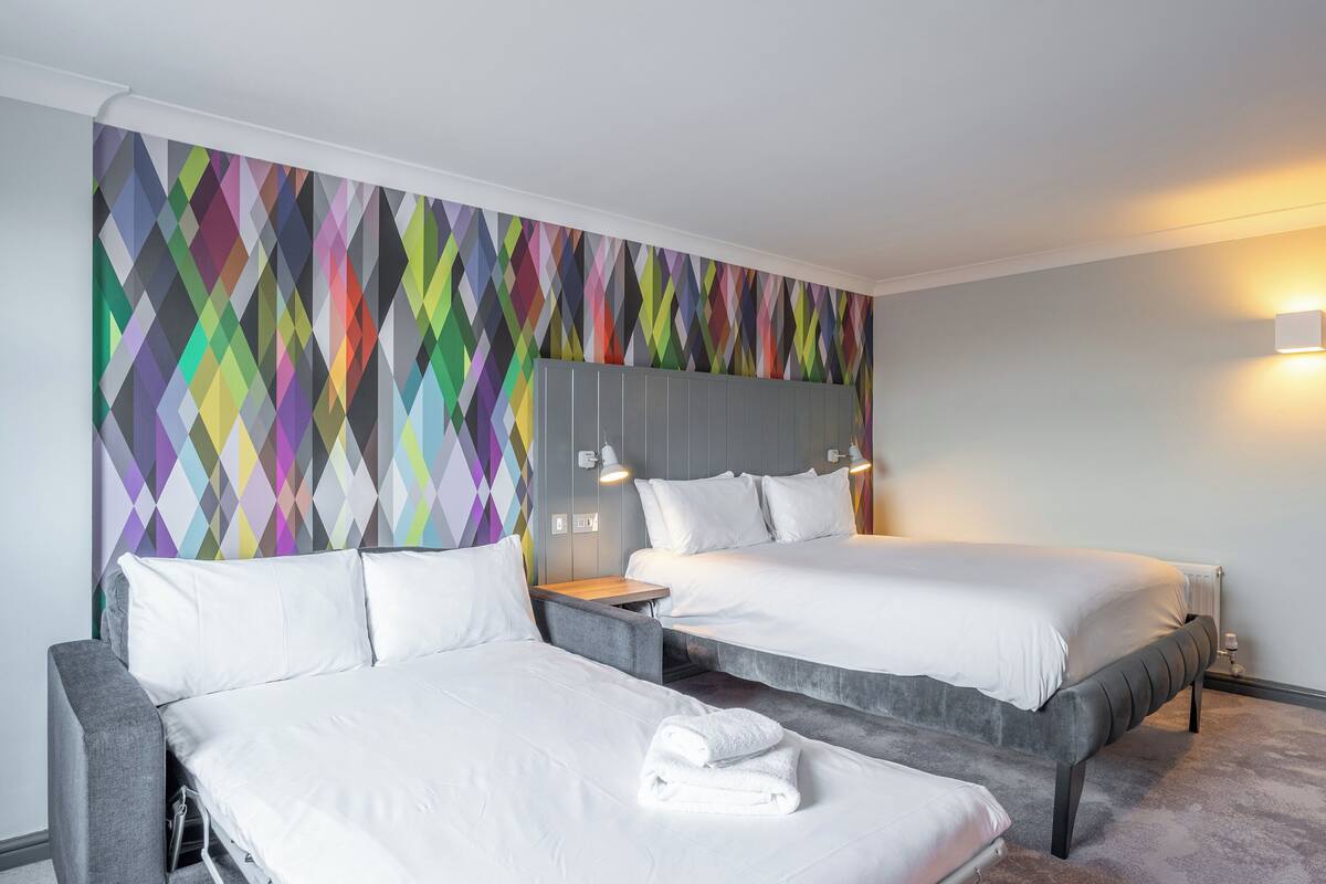 Habitación de hotel con dos camas, un muro de diseño geométrico y colores vivos, y una estética moderna.