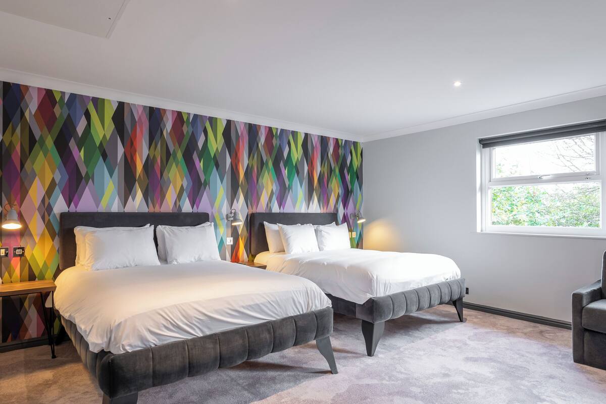 Un dormitorio moderno con dos camas, un diseño geométrico colorido en la pared y un ventanal que proporciona luz natural.