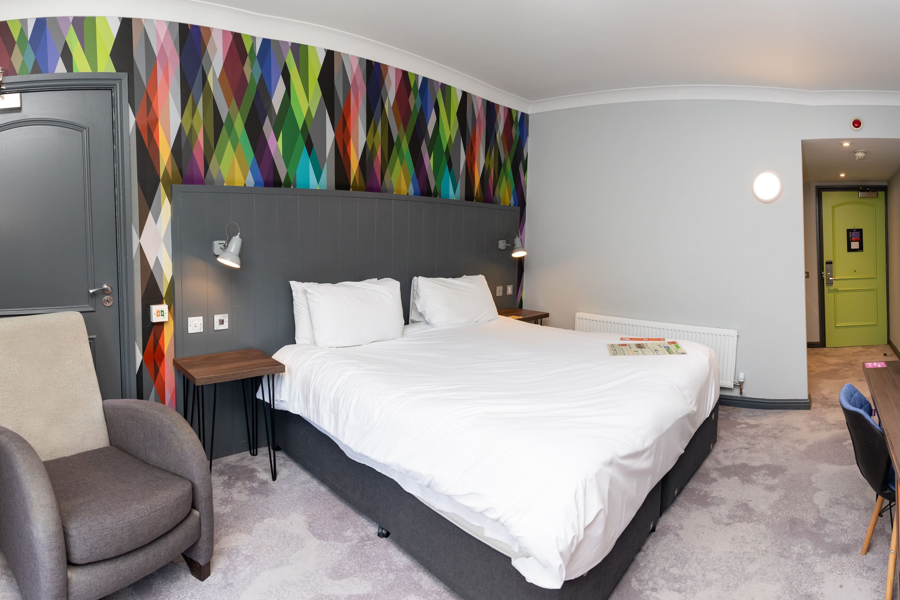 Een moderne hotelkamer met een groot bed, een grijze hoofdbord, een kleurrijke muurschildering, een grijze fauteuil en een klein tafeltje.