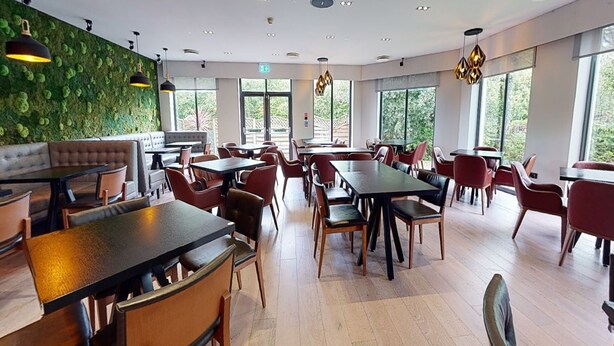 A modern restaurant with wooden tables and chairs, a green wall, and large windows. - Village Hotel St David's, Nr Chester (Deeside)