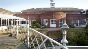 A brick building with a clock tower, a covered walkway, and a small pond. - Village Hotel St David's, Nr Chester (Deeside)