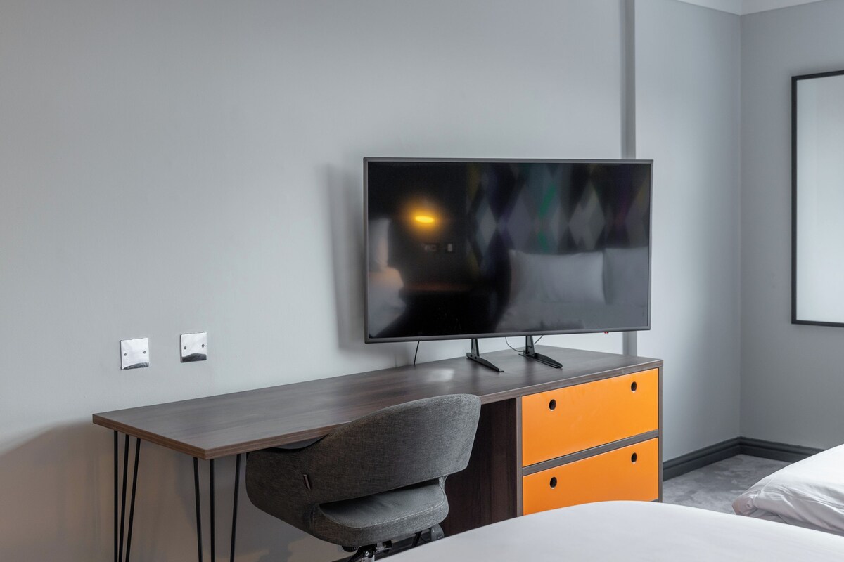 Une chambre d’hôtel moderne dotée d’un téléviseur à écran plat fixé au mur, d’un bureau en bois avec une chaise et d’un lit avec une literie blanche.