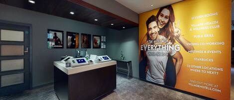 A hotel reception area with a large poster advertising various amenities such as stylish rooms, a village health and wellness club, a pub and grill, and a Starbucks.