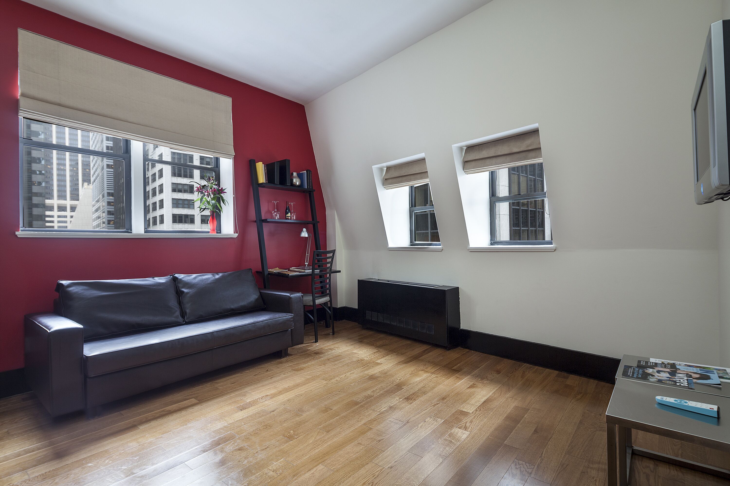 A living room with a black leather sofa, a wooden floor, a red accent wall, and a flat-screen TV mounted on the wall.