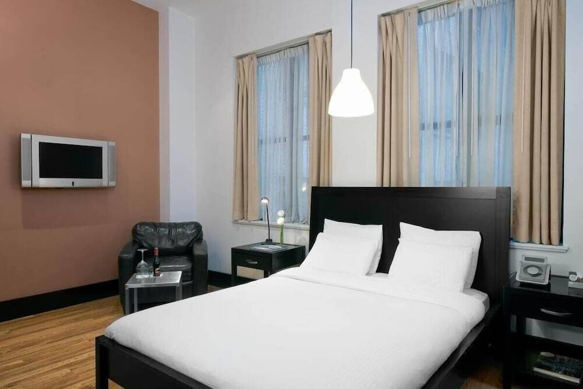 Habitación de hotel con cama, televisión, silla, mesita de noche y ventana con cortinas.