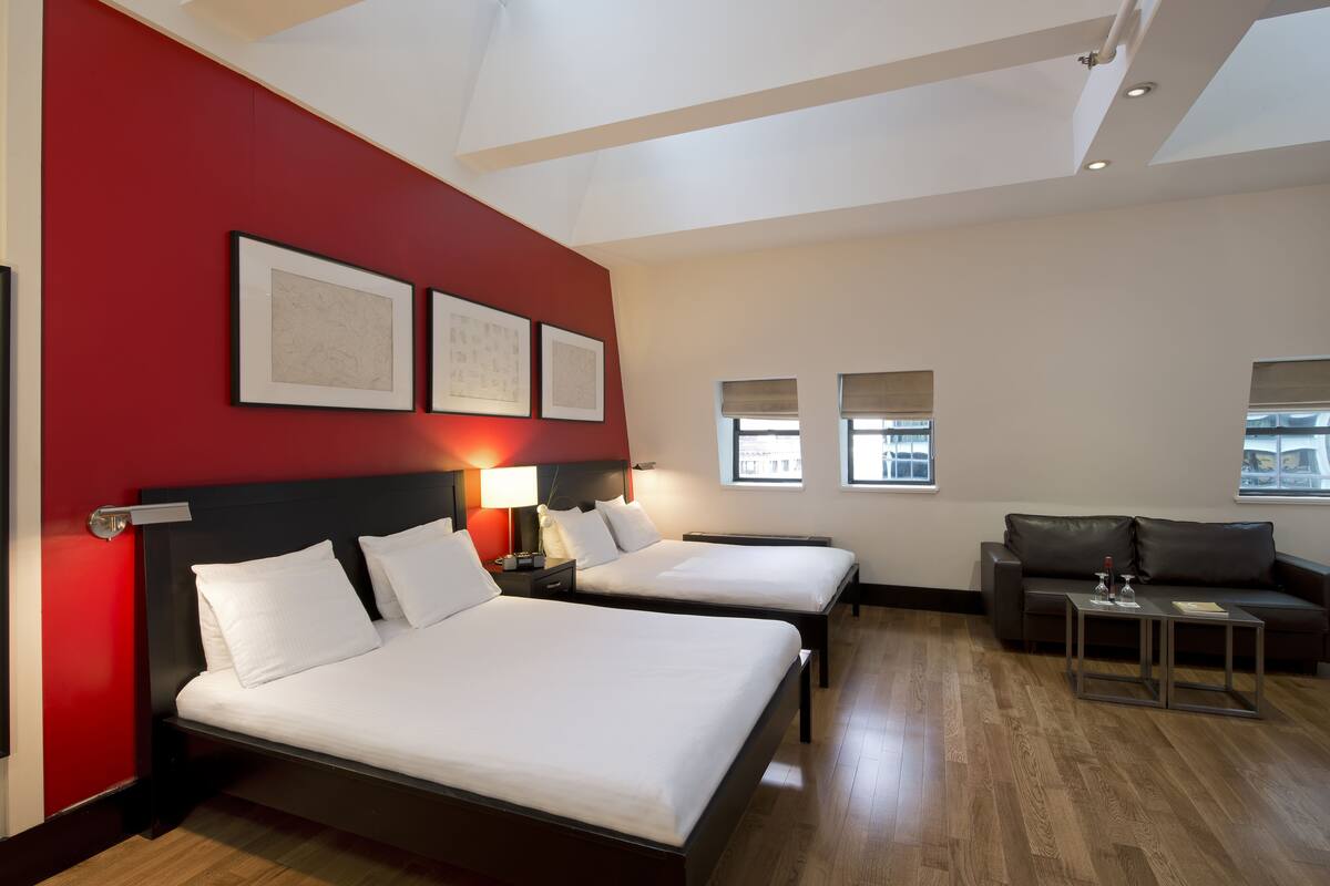 Habitación de hotel con dos camas, una pared de acento roja, obras de arte enmarcadas, un sofá y una mesita.